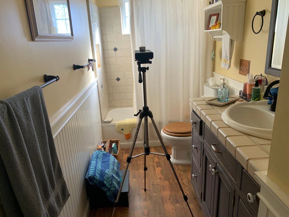 Bathroom interior with camera on tripod, vanity sink, toilet, towel rack, and striped bag on wooden floor