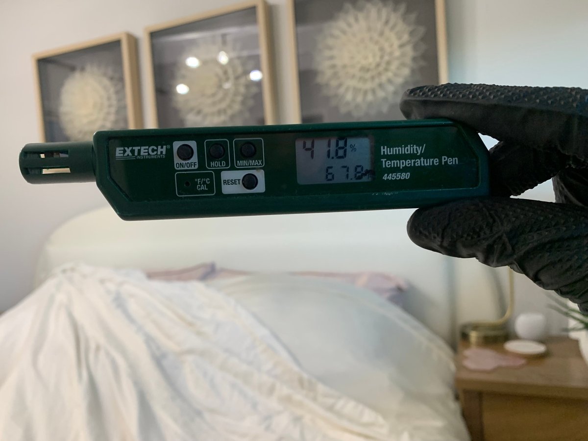 Hand holding an Extech digital humidity and temperature pen displaying readings of 71.8 degrees and 61.2 percent humidity, with bedroom furniture blurred in background