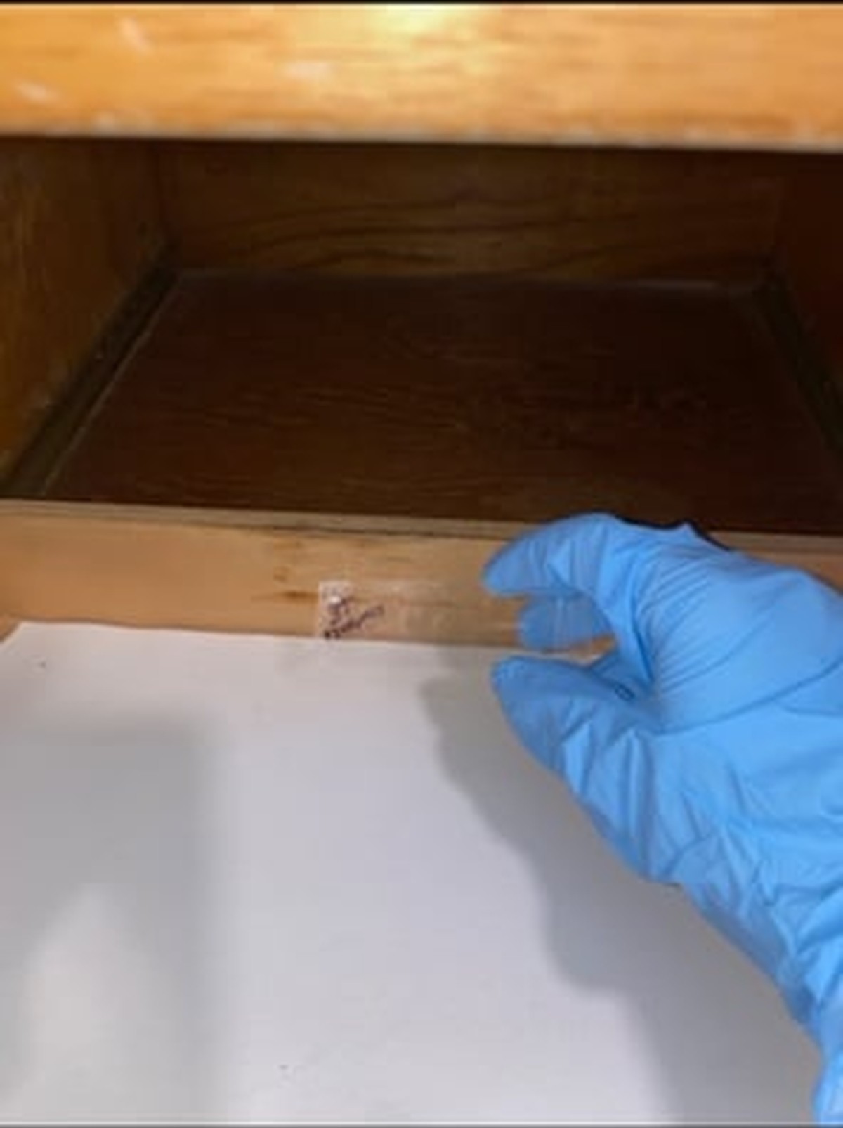 Hand in blue glove examining wooden drawer interior with dark stain, white cloth below