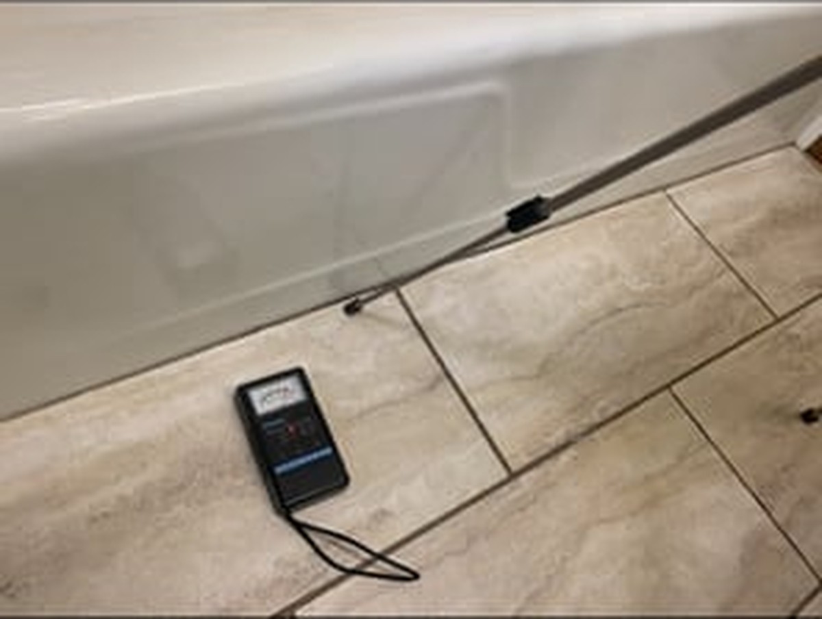 Digital moisture meter on tile floor next to white baseboard with black cables