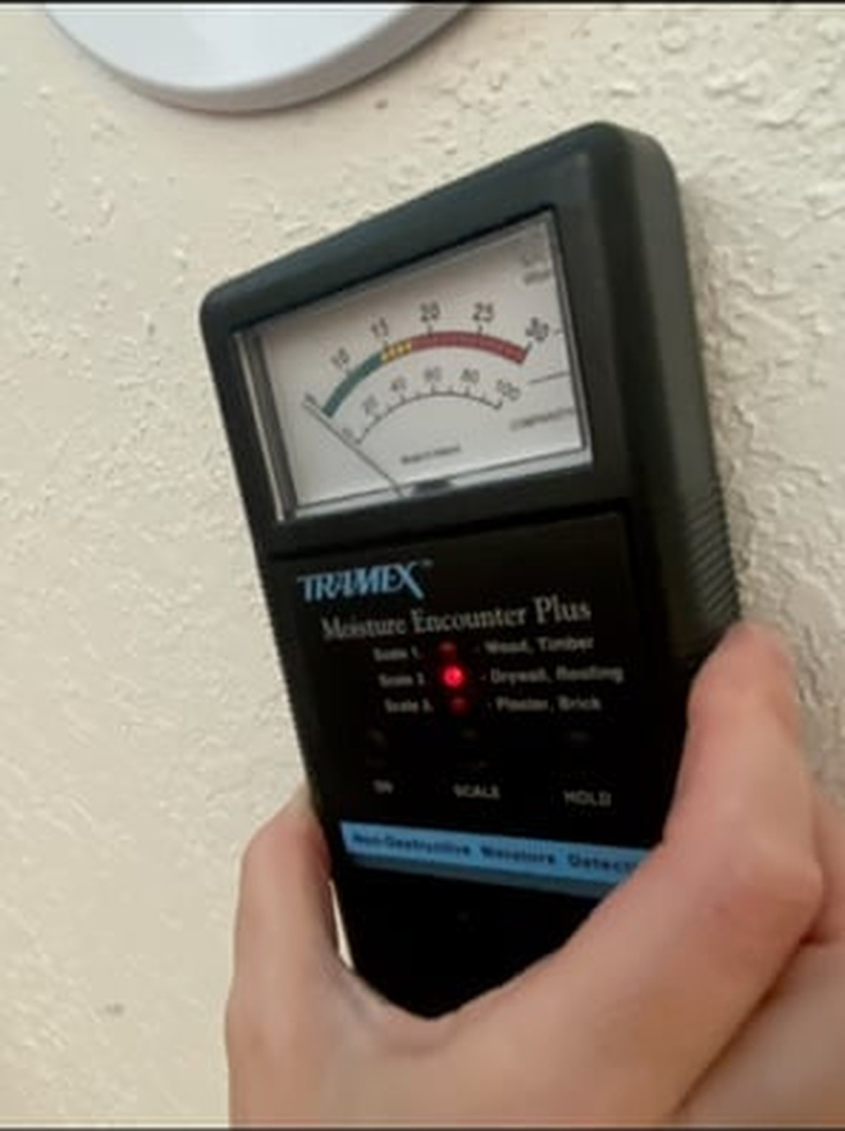 Hand holding a black Tramex moisture meter against a white textured wall surface during measurement